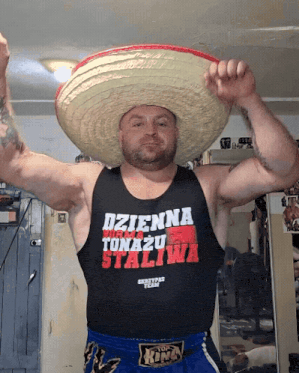 A man in a big straw sombrero and a black tank top dancing with his arms in the air